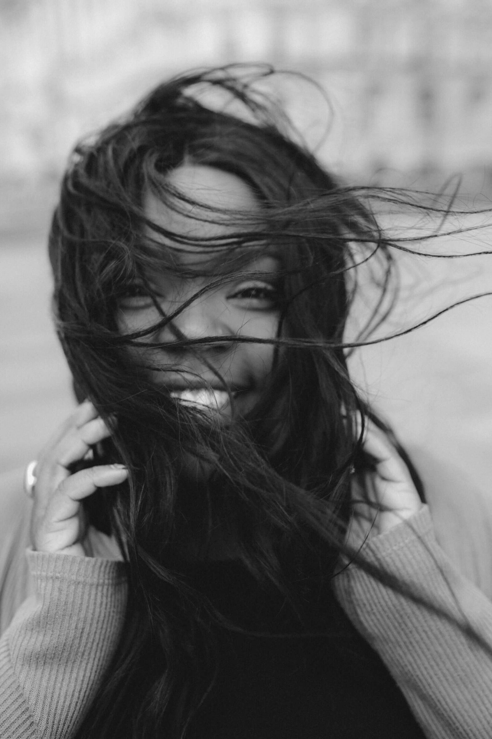 Portrait en noir et blanc d’une femme souriante, les cheveux emportés par le vent, vêtue d’un pull gris clair sur un haut noir.